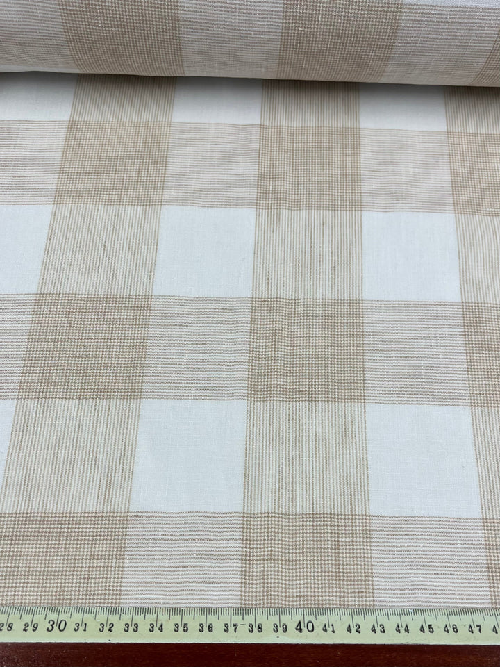 A roll of Pure Linen - Natural Check - 145cm by Super Cheap Fabrics featuring a beige and white plaid pattern is shown. Made from 100% linen, the fabric is laid out flat with a green ruler marked in centimeters along the bottom edge, showing a measurement of up to 55 centimeters—perfect for making outfits.