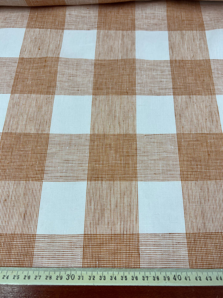 A lightweight fabric with a checkered pattern of orange and white squares. The orange squares contain diagonal lines creating a textured appearance, while the white squares are solid. At the bottom of the image is a green ruler showing measurements in centimeters. The fabric is called "Pure Linen - Copper Check - 145cm" by Super Cheap Fabrics.