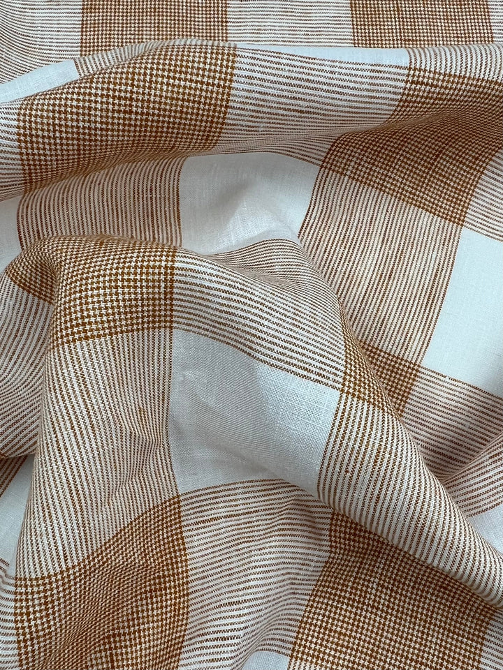 A close-up image of Super Cheap Fabrics' Pure Linen - Copper Check - 145cm featuring a checkered pattern in shades of brown and off-white. The lightweight fabric appears to be draped, creating folds and shadows that add texture to the image. The checkered design consists of alternating large and small squares.