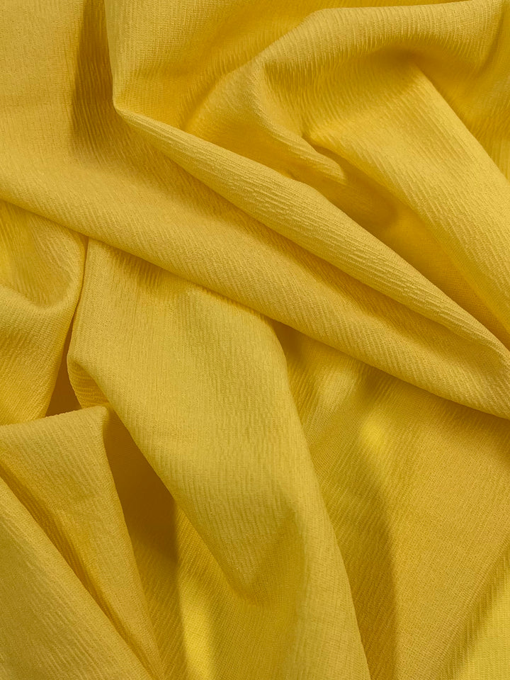 A close-up view of Super Cheap Fabrics’ Stretch Crinkle in Yellow – 140cm, featuring a vibrant polyester fabric with a soft, textured surface. Draped and folded to create gentle curves and shadows that add depth, embodying high fashion elements.