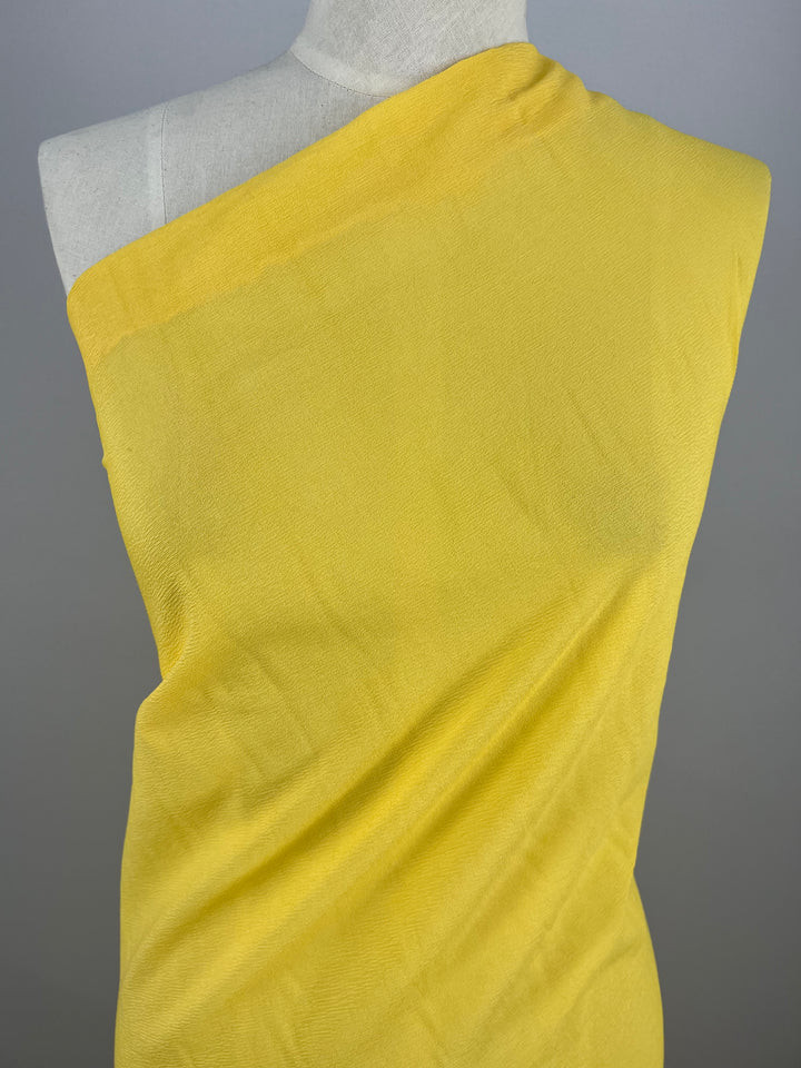 Against a neutral gray background, the Stretch Crinkle - Yellow - 140cm fabric from Super Cheap Fabrics is elegantly draped on a dress form mannequin in an asymmetric one-shoulder design, embodying high fashion.