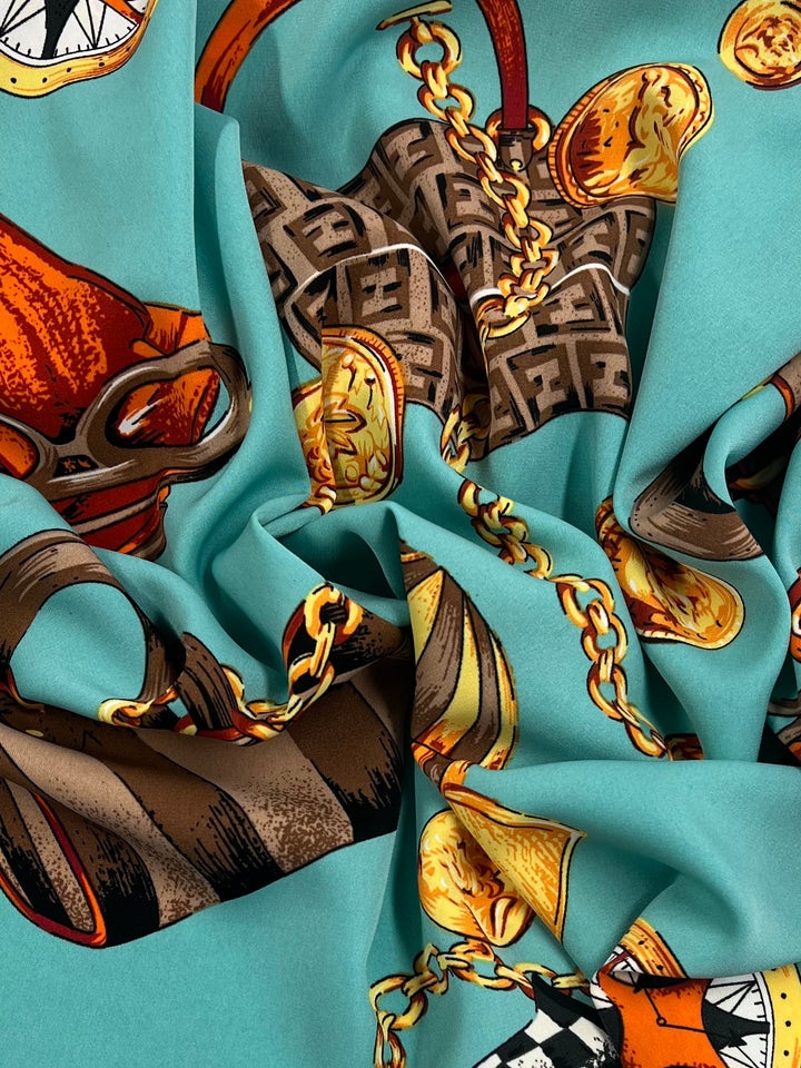 A close-up of Super Cheap Fabrics' Deluxe Print - Ocean Riche - 150cm featuring a pattern with twisted gold chains, ornate watches, and intricate geometric designs on a turquoise background. The artistic print showcases rich, detailed illustrations with a mix of warm and cool tones.