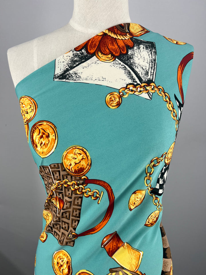 A mannequin draped in a teal Deluxe Print - Ocean Riche - 150cm fabric from Super Cheap Fabrics adorned with intricate gold chains, coins, and various colorful patterns including flowers and geometric shapes. The fabric covers one shoulder of the mannequin, showcasing the detailed print against a plain background.