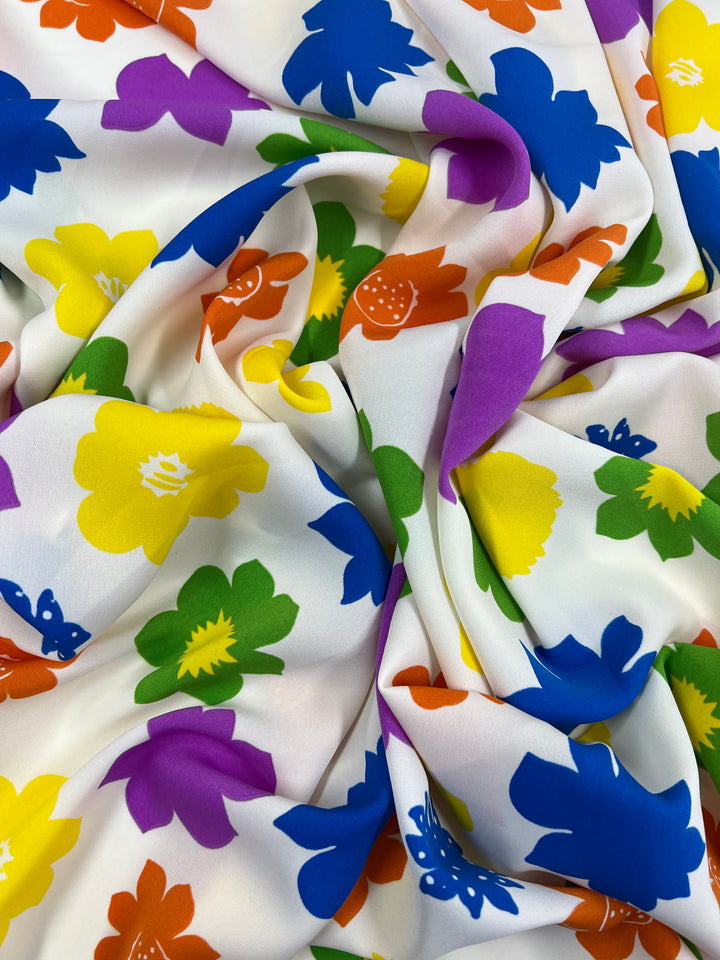 A close-up image of Deluxe Print - Flower Spread - 150cm draped and gathered, featuring a vibrant floral pattern. The flowers are in bold colors including blue, yellow, orange, green, and purple, set against a white background. The polyester fabric from Super Cheap Fabrics boasts a smooth and slightly glossy texture.