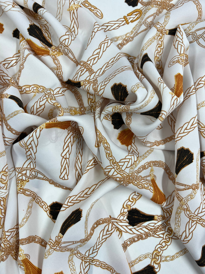 A close-up photo of Super Cheap Fabrics' Deluxe Print - Luxe Ties - 150cm white polyester fabric with an intricate gold chain and tassel pattern. The chains are braided and interconnected, with tassels in various colors, including black and gold, distributed throughout the design. The versatile textile is draped and slightly wrinkled.