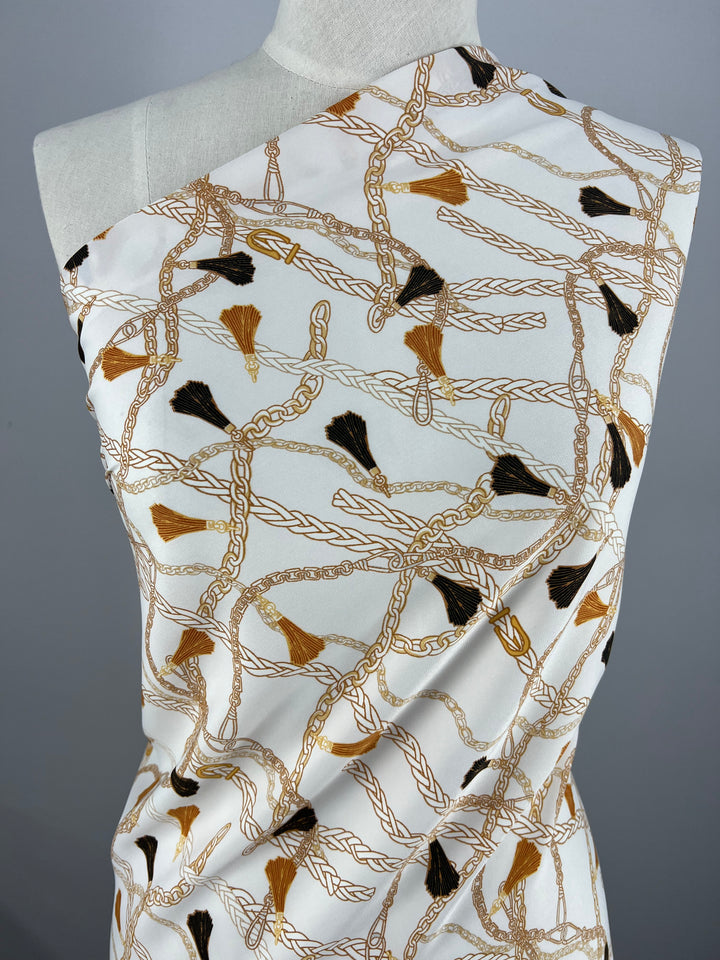 A piece of Super Cheap Fabrics' Deluxe Print - Luxe Ties - 150cm polyester fabric with a white background is draped over a mannequin. The fabric features an intricate pattern of gold chains and tassels in black and brown colors, exemplifying the refined elegance found in designer fabrics.