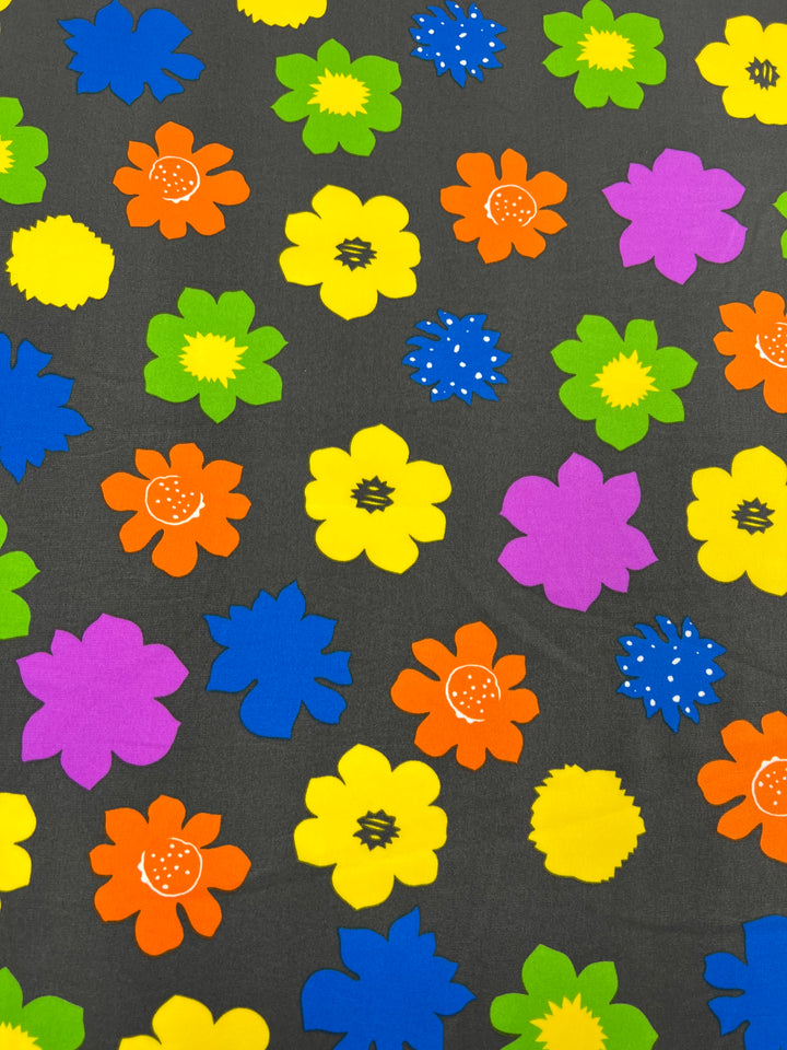 A designer fabric with a dark gray background and a vibrant pattern of colorful, stylized flowers. The flowers are in various bright colors including orange, yellow, purple, green, and blue, creating a lively and cheerful design. This versatile fabric is perfect for adding a pop of color to any project. Introducing the Deluxe Print - Flower Spread Retro - 150cm from Super Cheap Fabrics!