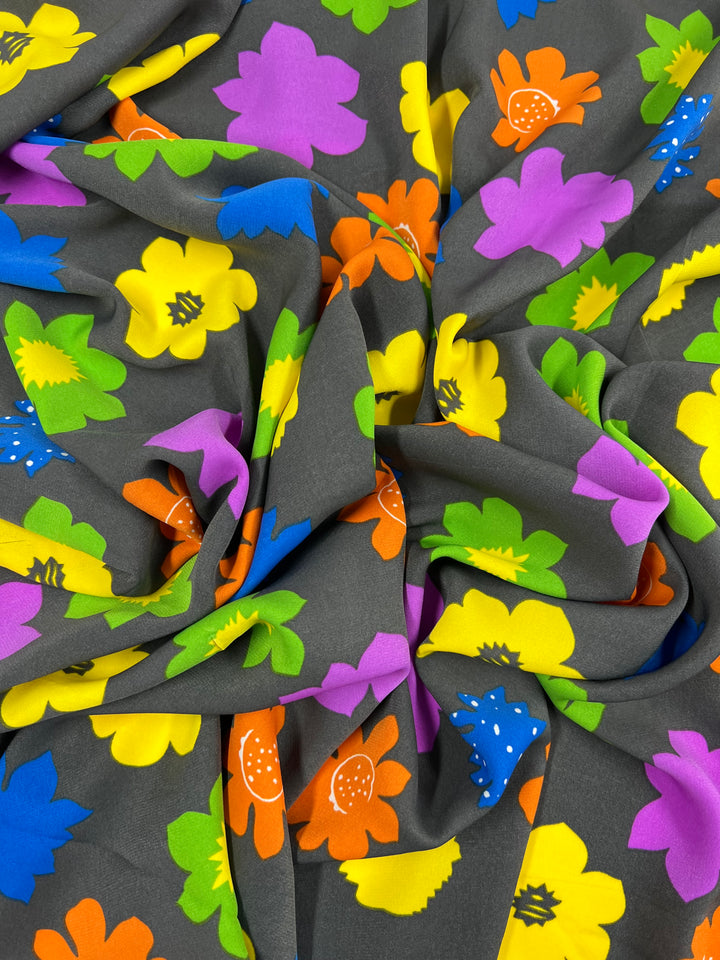 A close-up of a crumpled designer fabric featuring a vibrant floral pattern. The flowers are large, with colors including purple, yellow, green, orange, and blue. The background is dark gray, providing a striking contrast to the bright and colorful blooms. This is the Deluxe Print - Flower Spread Retro - 150cm from Super Cheap Fabrics.