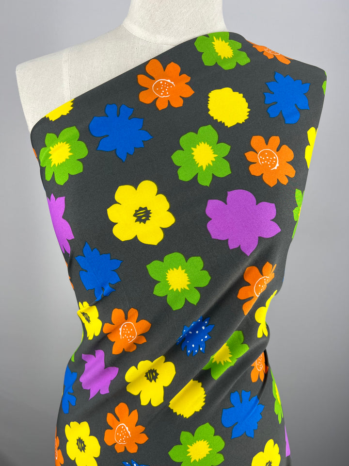 A mannequin draped with a dark fabric featuring a vibrant floral pattern. The flowers, in shades of orange, yellow, blue, green, and purple, are scattered across the polyester fabric of the Deluxe Print - Flower Spread Retro - 150cm by Super Cheap Fabrics, creating a lively and colorful design. The mannequin is displayed against a plain gray background.