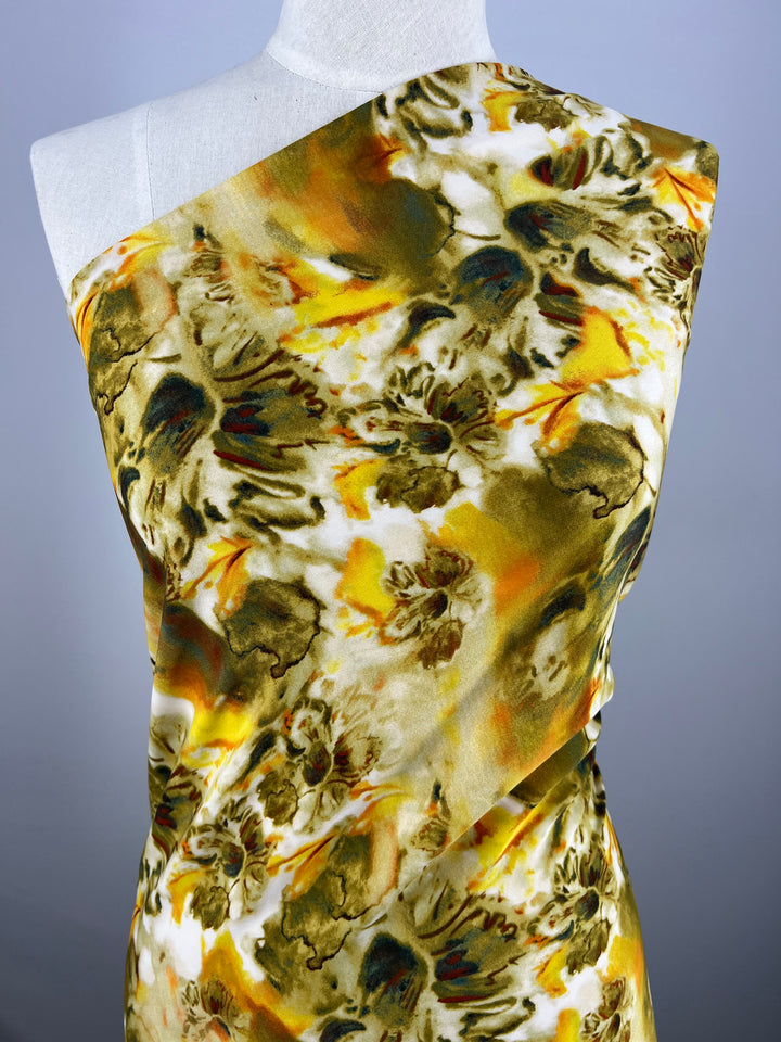 A mannequin draped in a colorful Deluxe Print - Flor Ray - 150cm with a vibrant abstract floral pattern. The designer fabric from Super Cheap Fabrics features shades of green, yellow, white, and brown, creating a lively and dynamic design. The background is a neutral gray, emphasizing the bold colors of this versatile fabric.