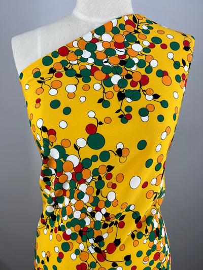 A sleeveless dress with a bright yellow background features an abstract pattern of overlapping circles in white, green, orange, and black. Made from versatile polyester fabric by Super Cheap Fabrics, the Deluxe Print - Yellow Dots & Vines - 150cm dress has an asymmetrical one-shoulder design. The lightweight material drapes elegantly on a mannequin.