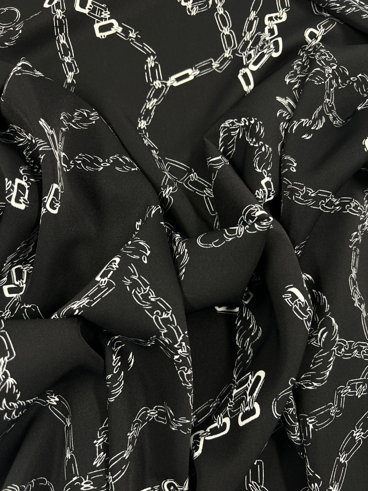 A close-up of a black Deluxe Print - MonoLinks - 150cm designer fabric by Super Cheap Fabrics featuring a pattern of white connected chains. The versatile fabric appears gathered and folded, creating shadows and depth, emphasizing the intricate chain design.