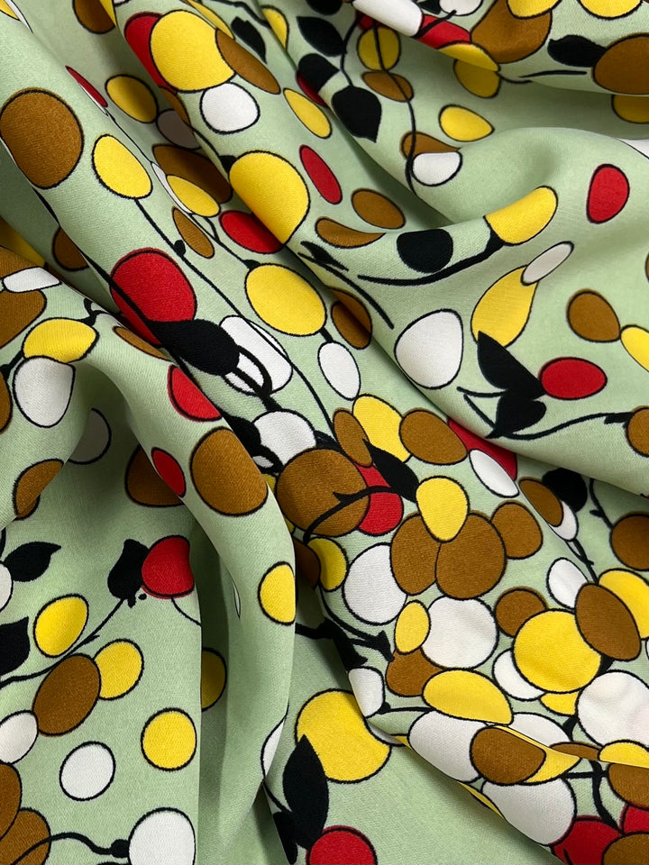 A close-up of a fabric with a vibrant pattern featuring clusters of multicolored circles in yellow, red, brown, and white on a light green background. The lightweight fabric appears to be gathered and folded, creating shadows and texture. This is the Deluxe Print - Sienna Dots & Vines - 150cm by Super Cheap Fabrics.