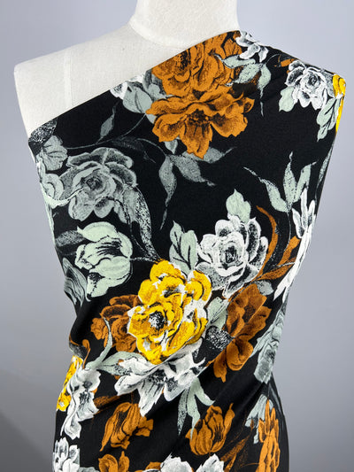 A mannequin displays the Super Cheap Fabrics' "Printed Crepe - Marigoldine - 150cm", a lightweight polyester with a bold floral pattern in orange, white, and gray on black, elegantly draped in a high-fashion one-shoulder style.