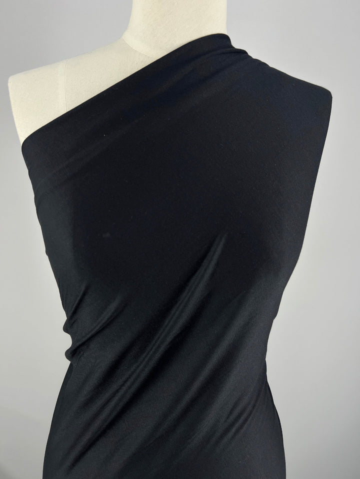 A mannequin dressed in a sleek, one-shoulder black garment made of Super Cheap Fabrics' Cotton Jersey - Black - 150cm. The background is a plain, light gray color, highlighting the elegant simplicity of the outfit.