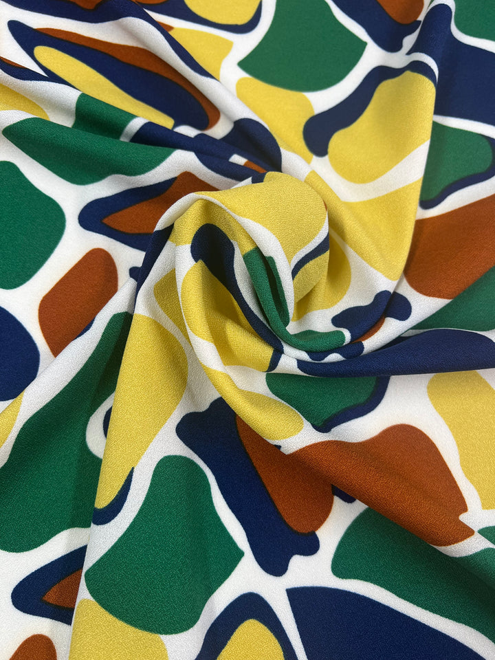 A close-up of Super Cheap Fabrics' Printed Crepe - Camo, a lightweight polyester with a vibrant abstract pattern in green, yellow, blue, brown, and white. The high fashion fabric is artfully twisted at the center for gentle folds and dynamic texture. Width: 150cm.