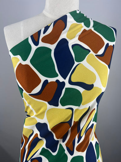 A mannequin wears the Super Cheap Fabrics' Printed Crepe - Camo, a sleek one-shoulder style in lightweight polyester. This fabric features vibrant abstract shapes in green, orange, brown, yellow, and blue on a white background, perfect for high fashion statements. Width: 150cm.