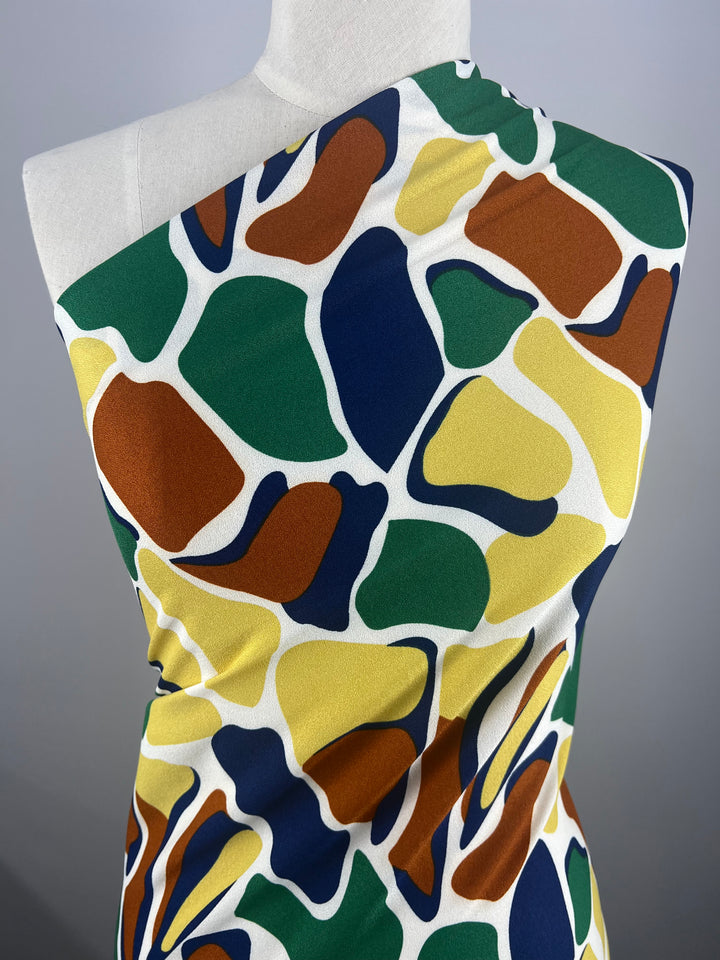 A mannequin wears the Super Cheap Fabrics' Printed Crepe - Camo, a sleek one-shoulder style in lightweight polyester. This fabric features vibrant abstract shapes in green, orange, brown, yellow, and blue on a white background, perfect for high fashion statements. Width: 150cm.