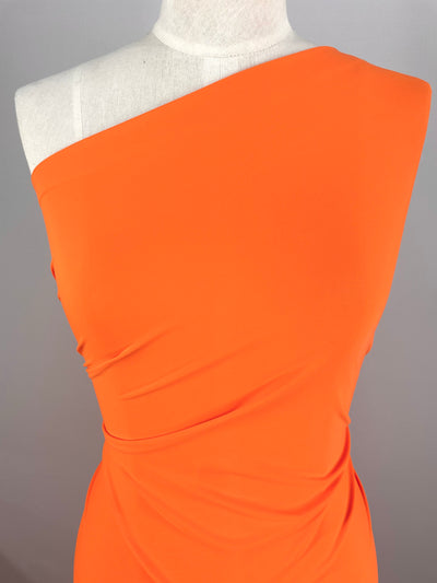 A mannequin is dressed in a vibrant, exotic orange one-shoulder dress made from Super Cheap Fabrics' Nylon Lycra. The material, which measures 150cm wide, is ruched at the sides to create a draped effect. The plain grey background enhances the bright color and stylish design of the dress.