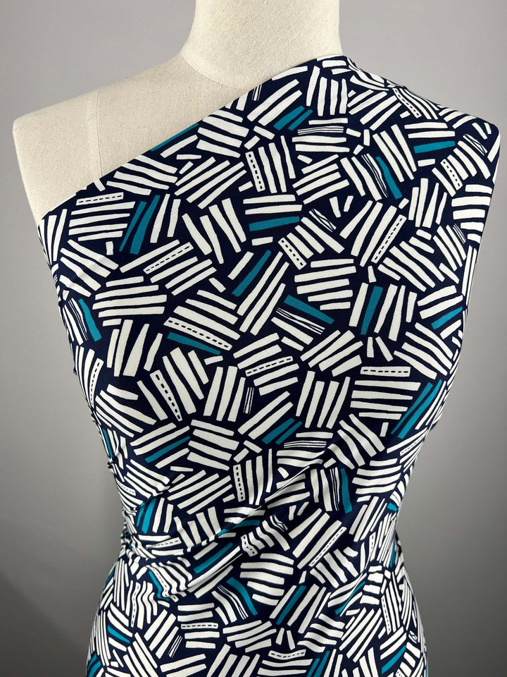 A mannequin displays the "Printed Lycra - Freehand - 147cm" by Super Cheap Fabrics: a one-shoulder dress in polyester/spandex, adorned with geometric white, navy, and teal stripes on black. The medium-weight fabric drapes smoothly, enhancing the asymmetrical design against a gray backdrop.