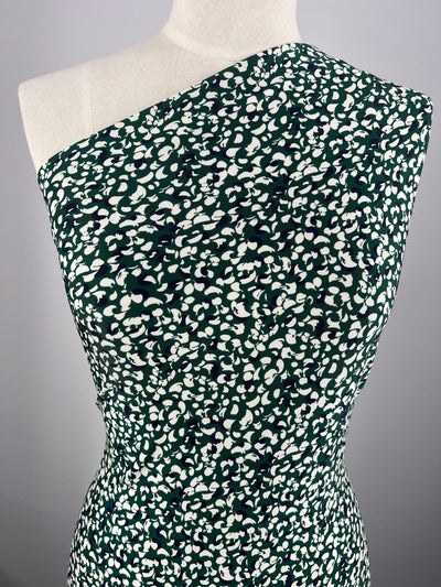 A mannequin showcases a one-shoulder dress made from Super Cheap Fabrics' Printed Lycra - Petals, featuring medium-weight Polyester/Spandex material adorned with a green and white abstract pattern. The modern, form-fitting design is beautifully highlighted against a plain grey background.