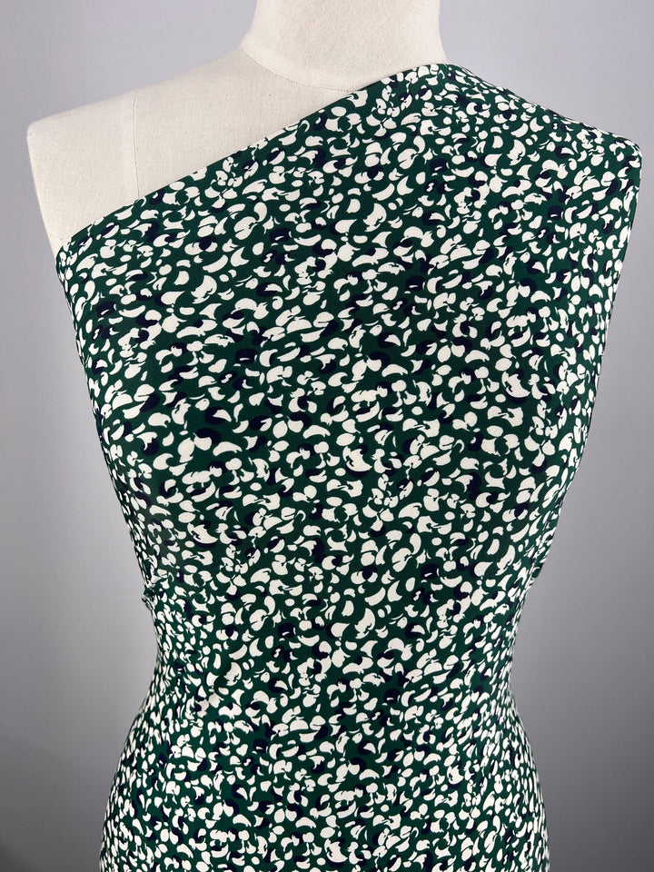 A mannequin showcases a one-shoulder dress made from Super Cheap Fabrics' Printed Lycra - Petals, featuring medium-weight Polyester/Spandex material adorned with a green and white abstract pattern. The modern, form-fitting design is beautifully highlighted against a plain grey background.