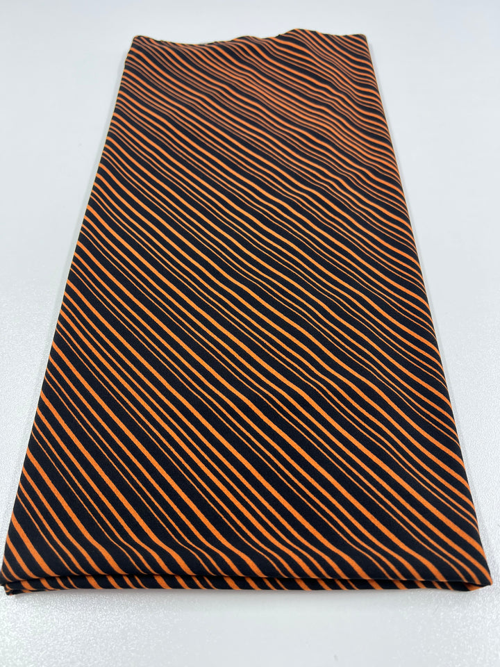 A neatly folded piece of REMNANT - Lycra - 90cm from Super Cheap Fabrics, featuring a pattern of diagonal orange and black stripes, is placed on a white surface. The dark and light stripes create a striking contrast, accentuating the geometric design of the fabric.