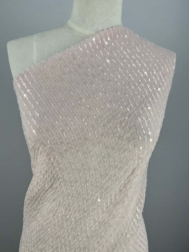 A dress form draped in **Evening Sequin - Baby Pink - 150cm** by **Super Cheap Fabrics**. The medium weight material has a textured, ribbed pattern and is adorned with small sparkling sequins, creating a shimmering effect. The background is plain gray, allowing the fabric details to stand out.