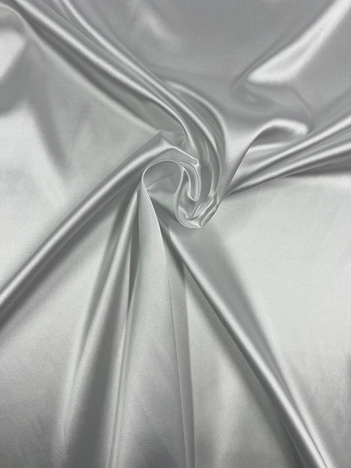 A close-up of the Satin Deluxe from Super Cheap Fabrics in white, showcasing its smooth, luxurious texture and soft sheen. The lightweight fabric is gently gathered in the center, creating subtle folds and a textured surface that highlights its shiny finish and glossy, reflective quality.