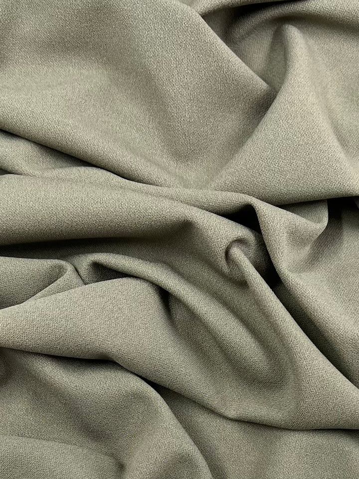 A close-up view of Super Cheap Fabrics' Plain Scuba Crepe in Laurel Oak, a light olive green material with a soft, matte texture. The stretchy fabric is displayed in gentle folds and creases, creating a subtle play of shadows and highlights on its 170cm wide surface, making it perfect for home decor projects.