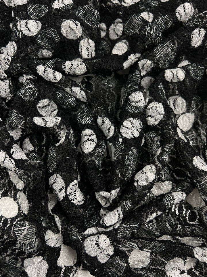 A close-up image of a black, sheer mesh fabric with swirling patterns, decorated with white polka dots. The delicate lace texture appears intricate, with the interplay of black and white creating a striking contrast. The fabric is "Evening Lace - Lemony - Black & White - 142cm" by Super Cheap Fabrics.