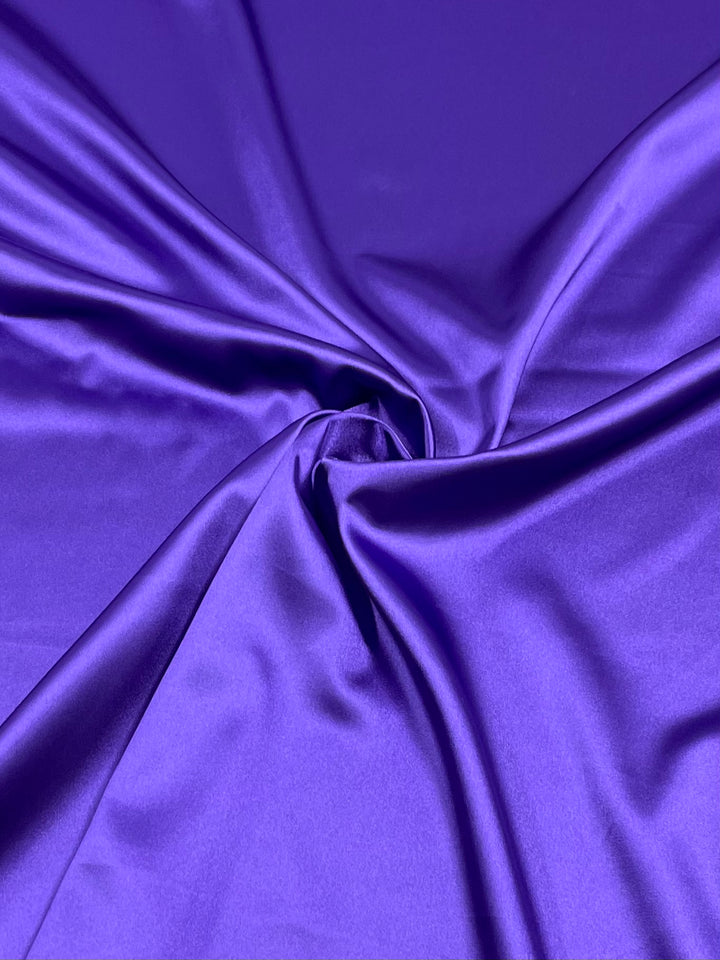 A close-up image of the smooth, shining Satin Deluxe - Purple - 150cm fabric from Super Cheap Fabrics. The lightweight material is gathered in gentle folds, reflecting light to showcase its lustrous texture and shiny finish.