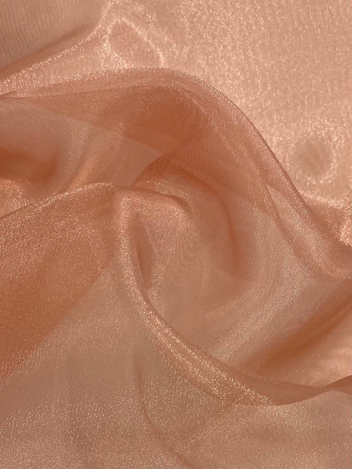 A close-up of a peach-colored sheer polyester fabric known as Organza - Shrimp, offered by Super Cheap Fabrics, features a glossy texture. This lightweight material is perfect for tops, showcasing soft crumples that reflect light and form delicate folds and shadows.